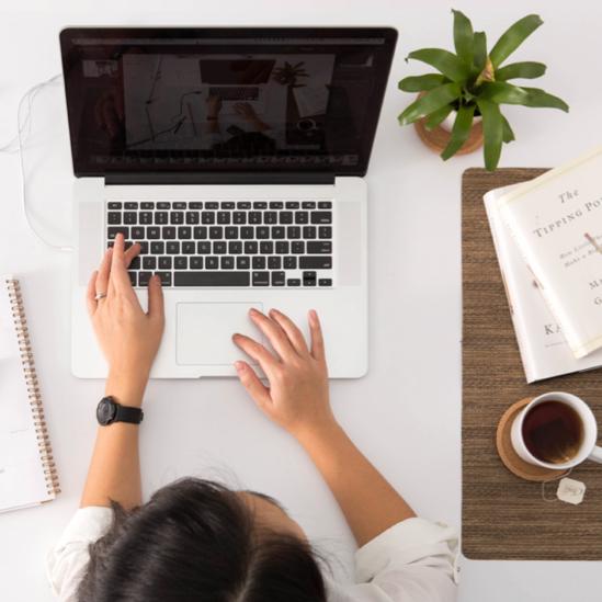 Brunette woman wearing a white shirt and black watch drinks tea at her table while using her MacBook.